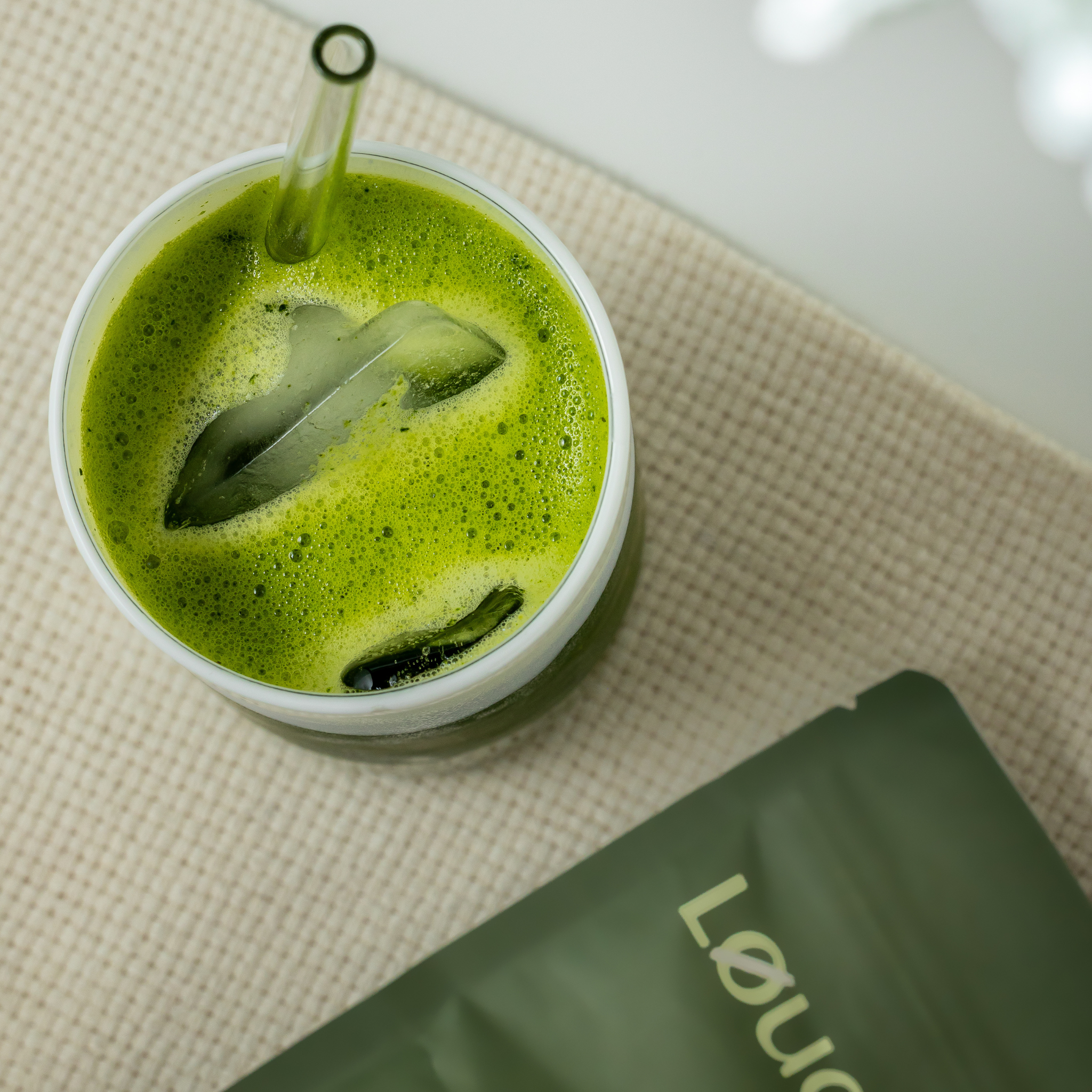 Løuco green protein smoothie in a cup with a straw on a textured surface, next to a Løuco Nourish package.