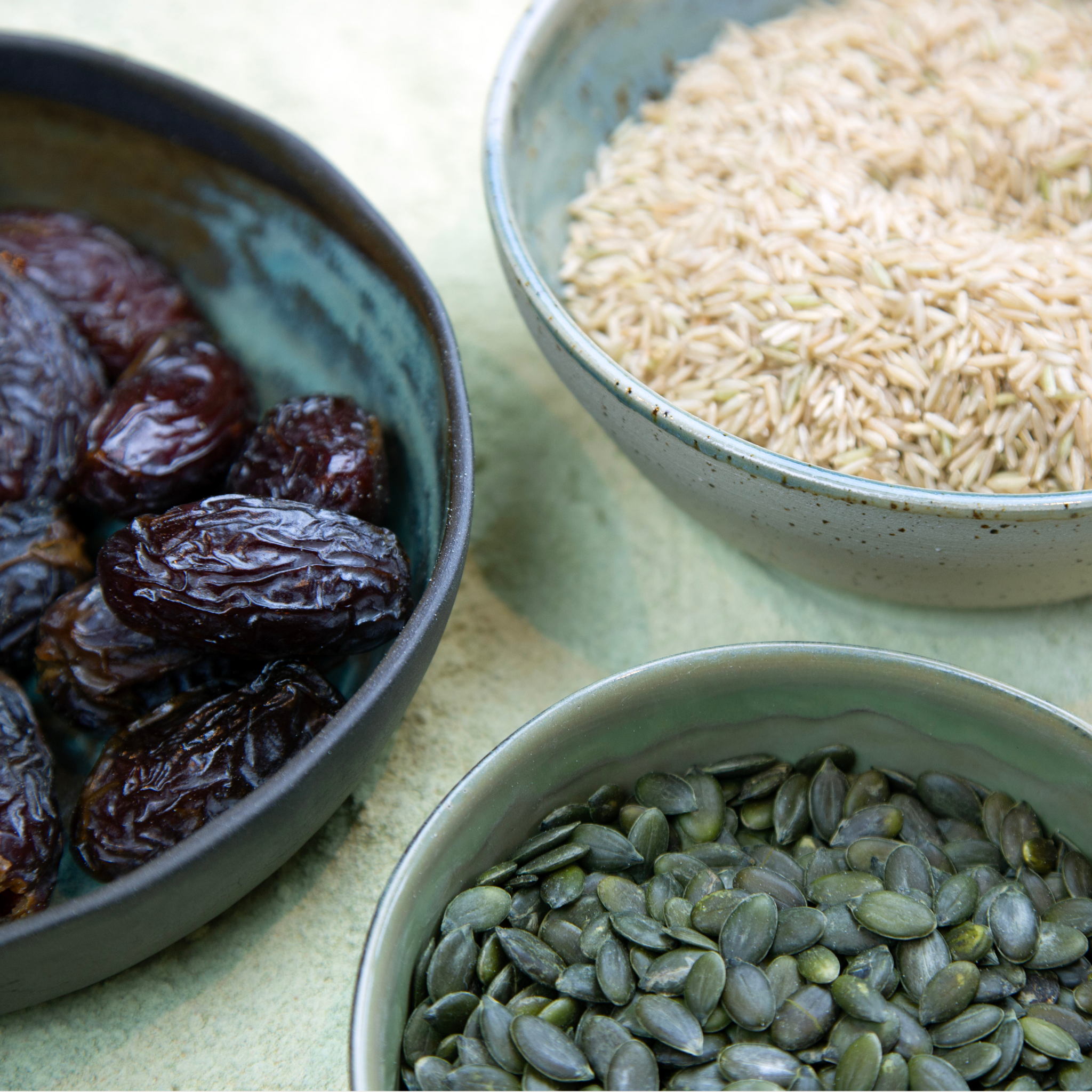 Three bowls containing dried dates, brown rice, and green pumpkin seeds on a light surface to show Løuco food-based ingredients. 