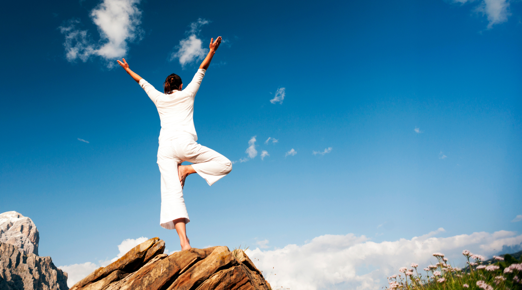 Person in white outfit standing on a rock with arms outstretched against a blue sky to represent LØUCO empowering values.
