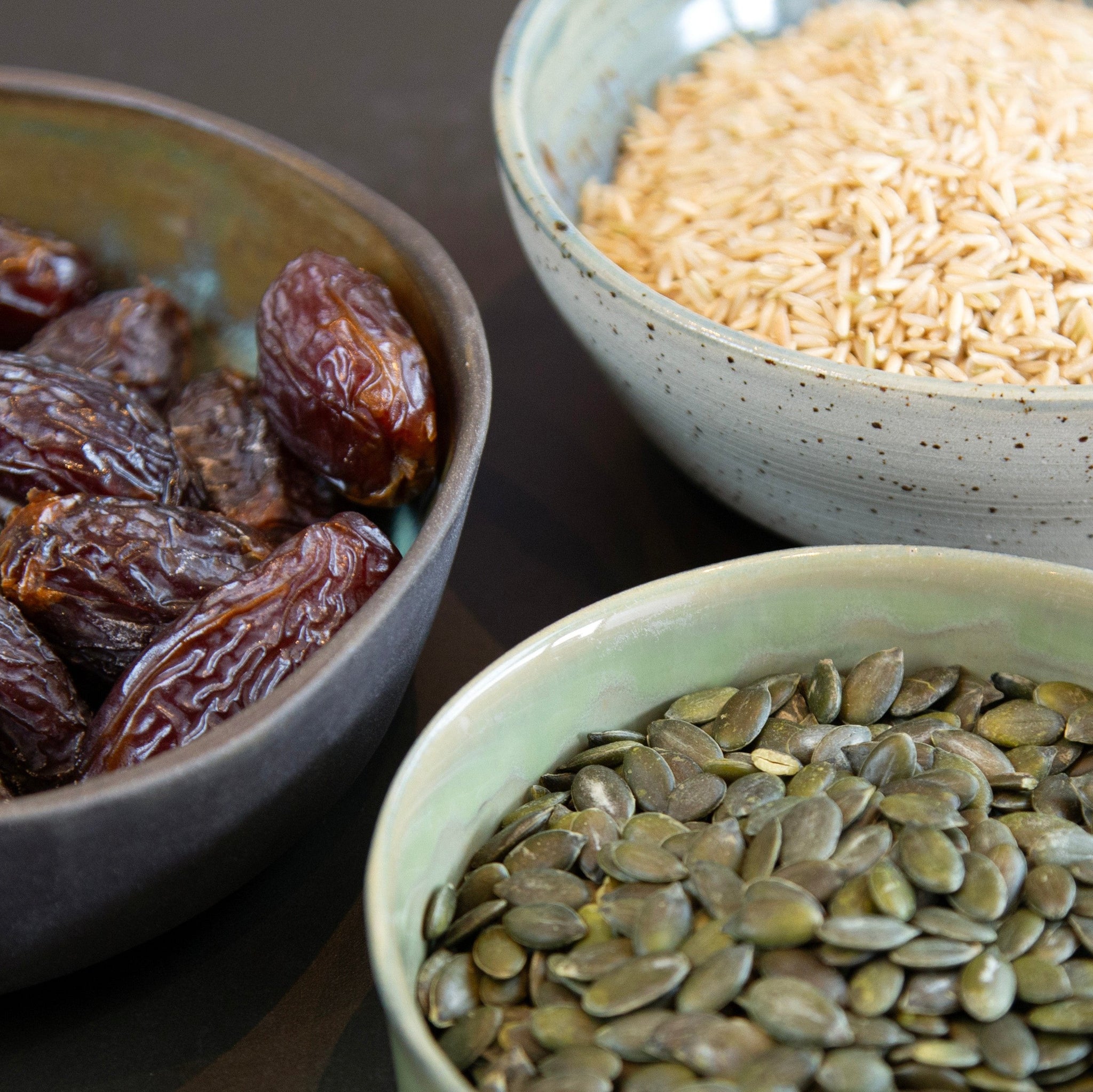 Three bowls of LØUCO protein powder ingredients containing dates, rice, and pumpkin seeds on a dark surface.